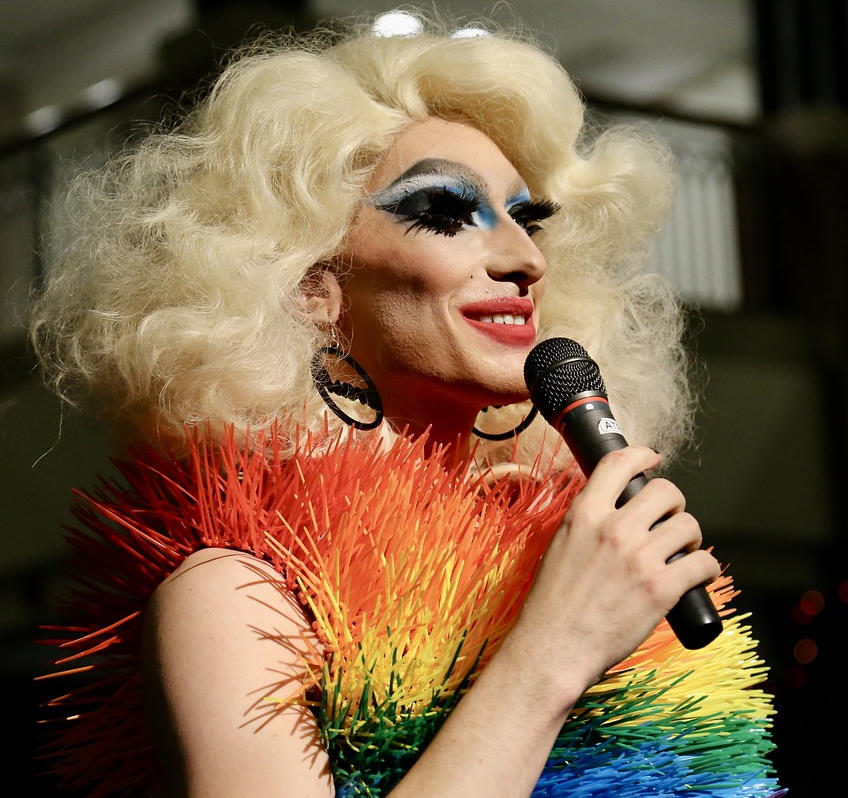 Eine Drag-Queen mit Mikrofon in der Hand und Regenbogenschal um den Hals.